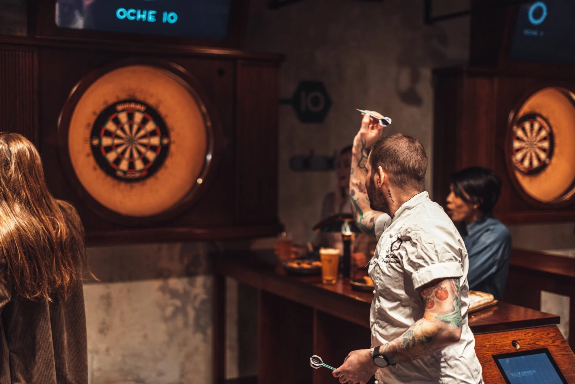 Mann i hvit kokkejakke kaster dartpil mot en dartskive, mens andre ser på ved et bord.