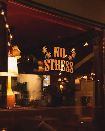 Vindu med oransje skrift «No Stress» og palmer, med lamper som lyser inne i baren om kvelden.